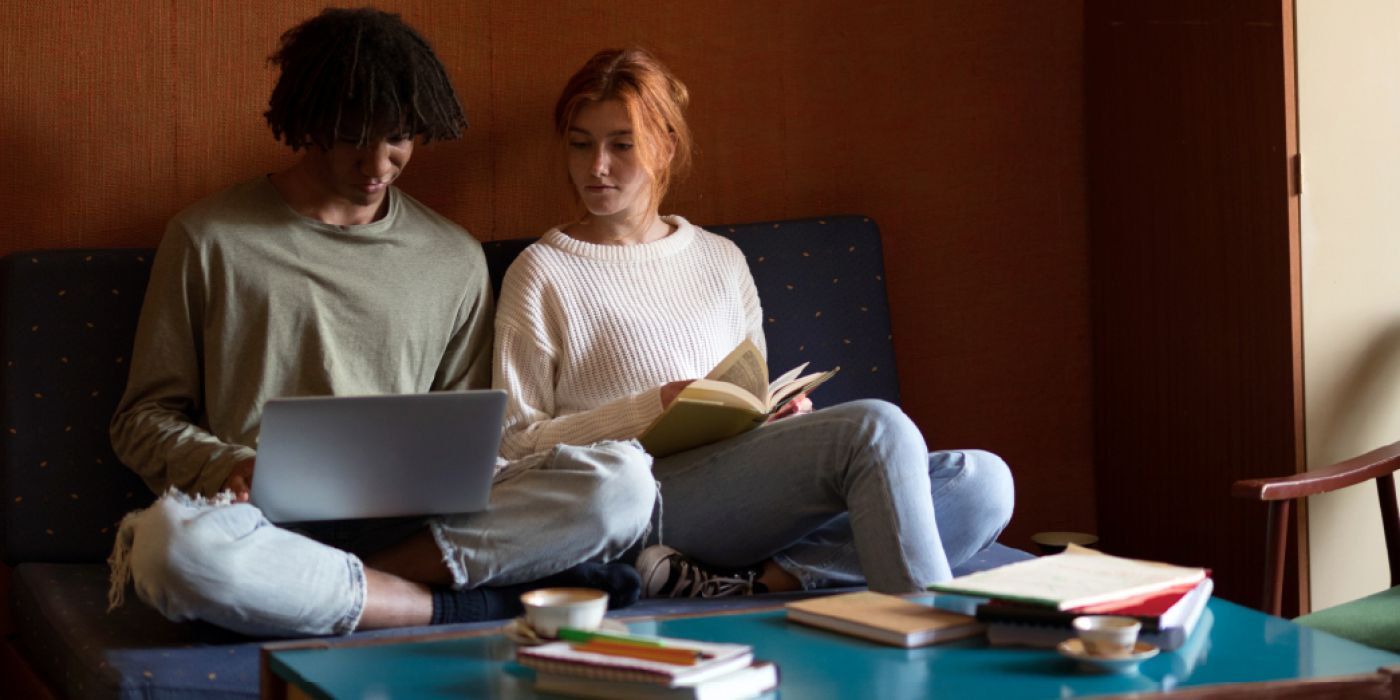 Two people sitting on a couch, using a laptop and reading a book, with a coffee table and books in front of them.
