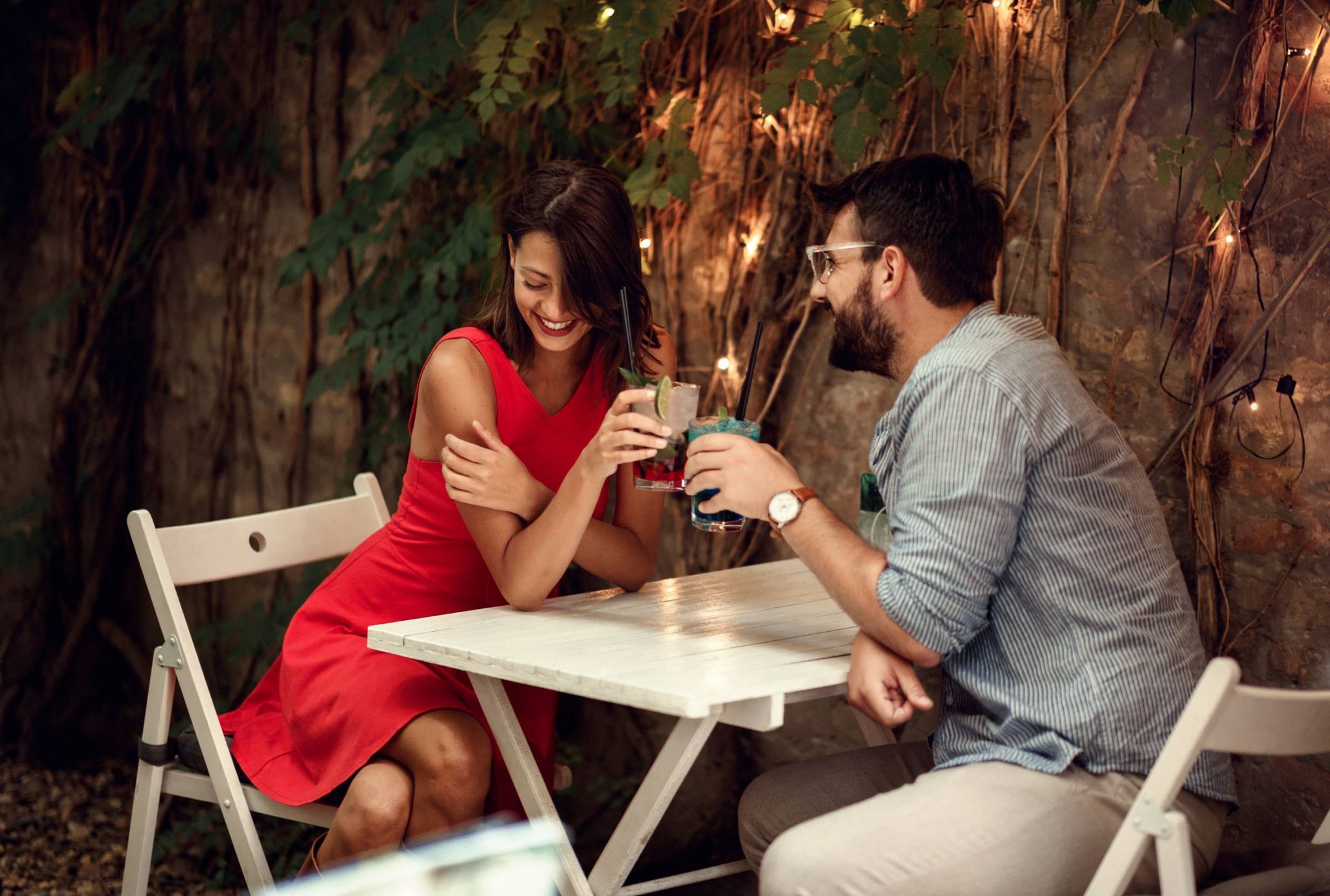A woman in a red dress and a man in a striped shirt enjoy drinks at an outdoor table with string lights and greenery.