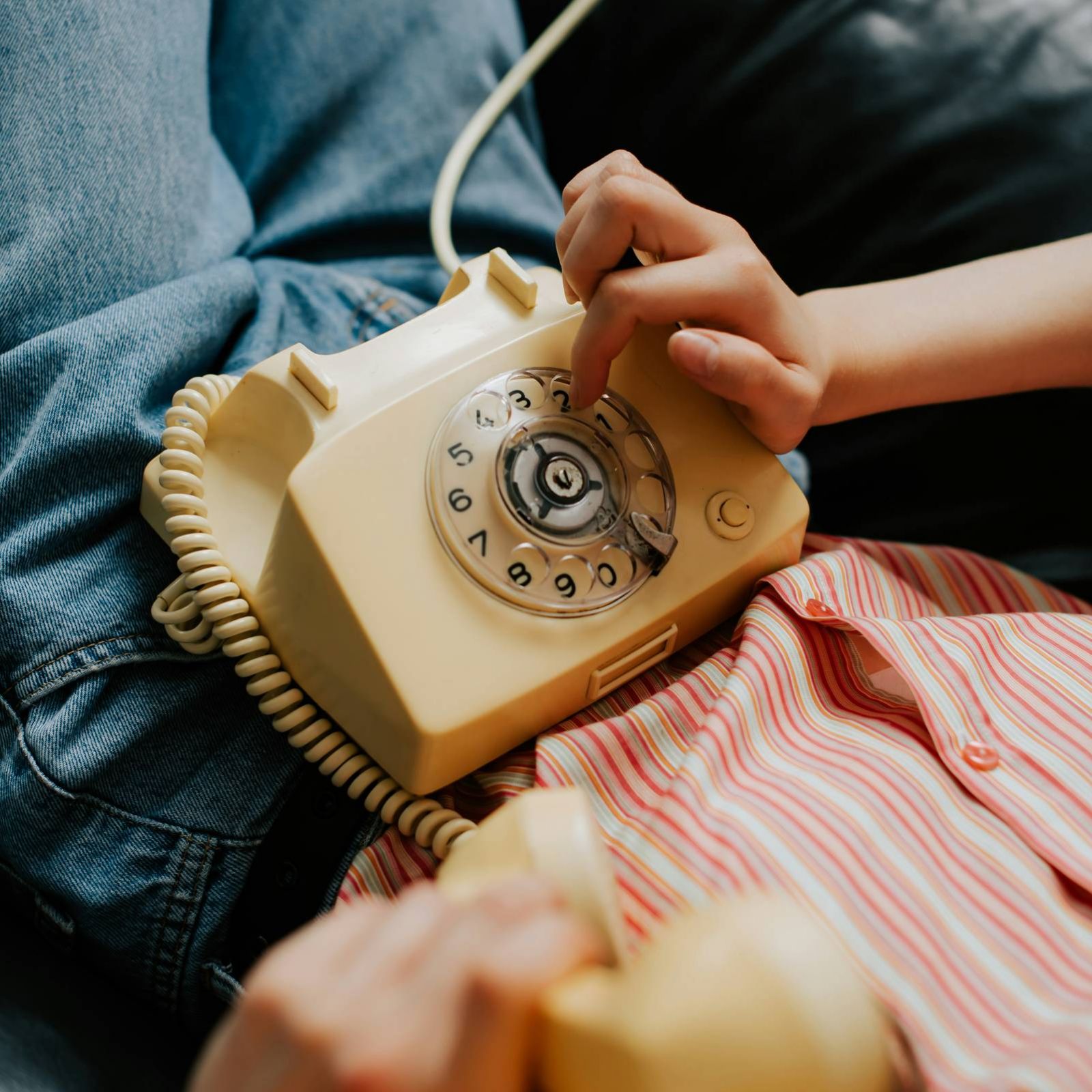 Person dialing a vintage rotary phone while holding the handset, wearing jeans and a striped shirt.