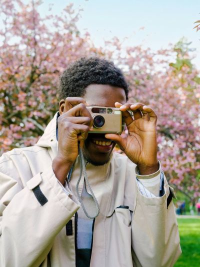 A person in a light coat smiles while taking a photo with a camera in front of blooming pink trees.
