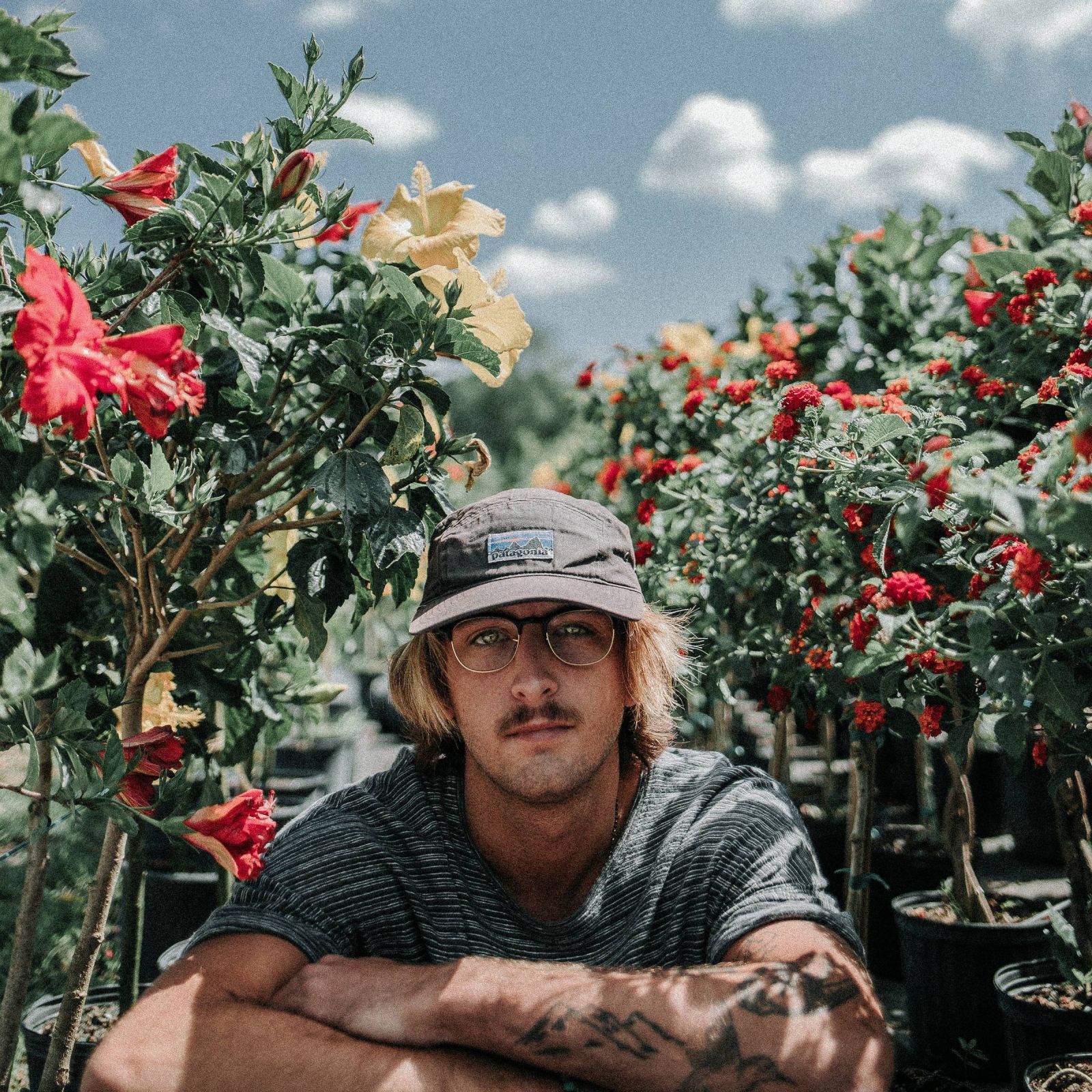 Person in glasses and a hat sits among colorful flowering plants under a blue sky.