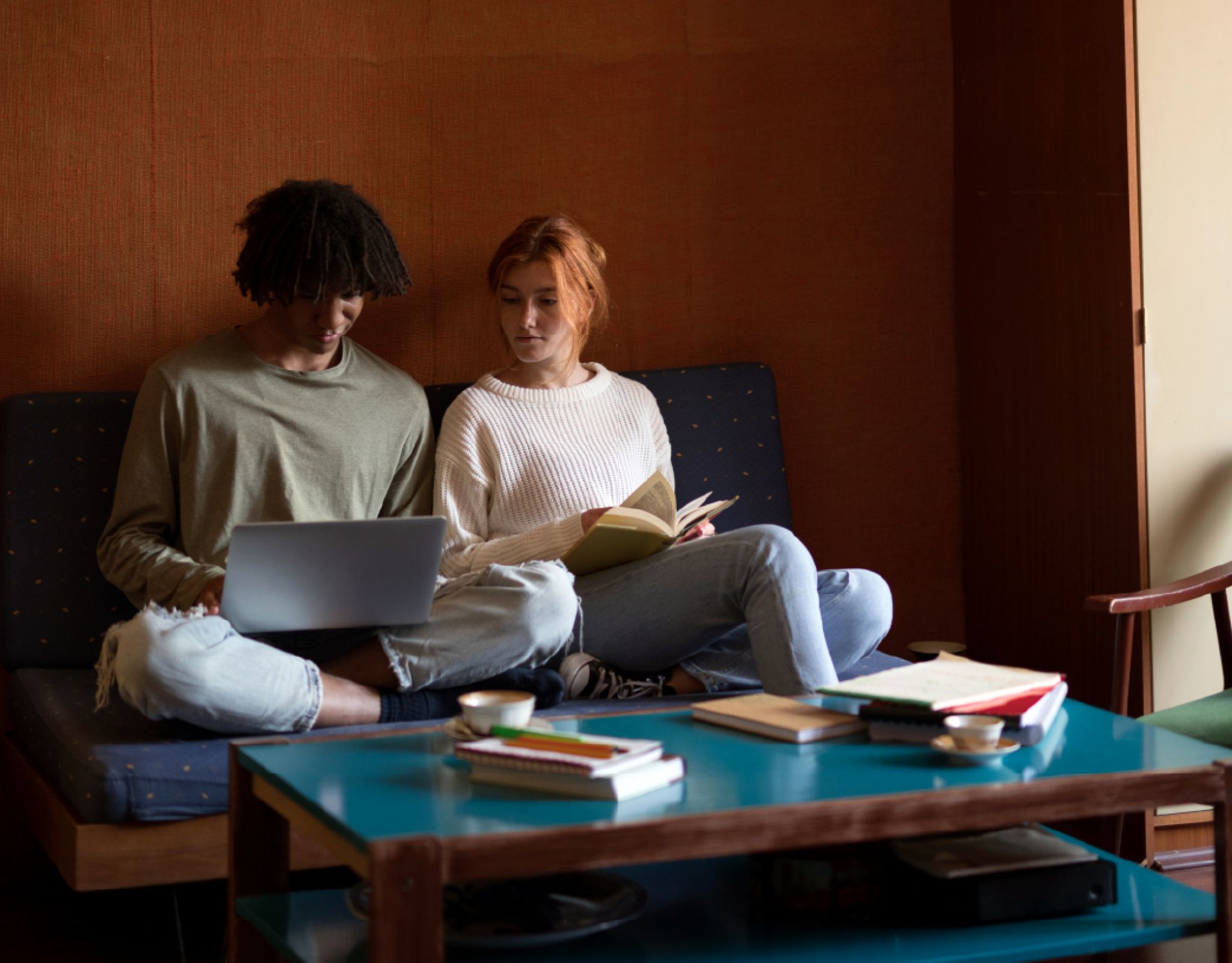 Two people sitting on a couch, using a laptop and reading a book, with a coffee table and books in front of them.