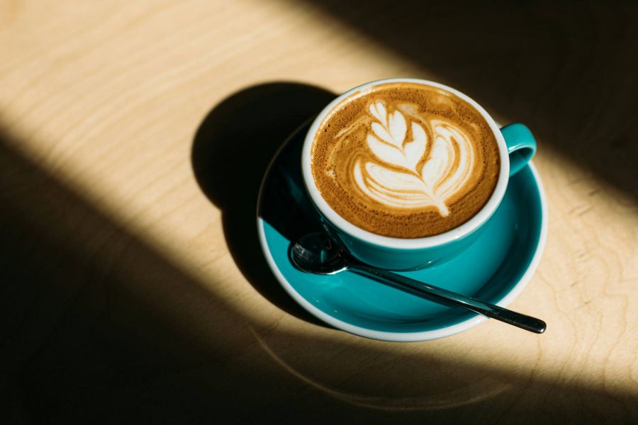 A teal cup of latte with leaf latte art, placed on a wooden table, lit by sunlight.