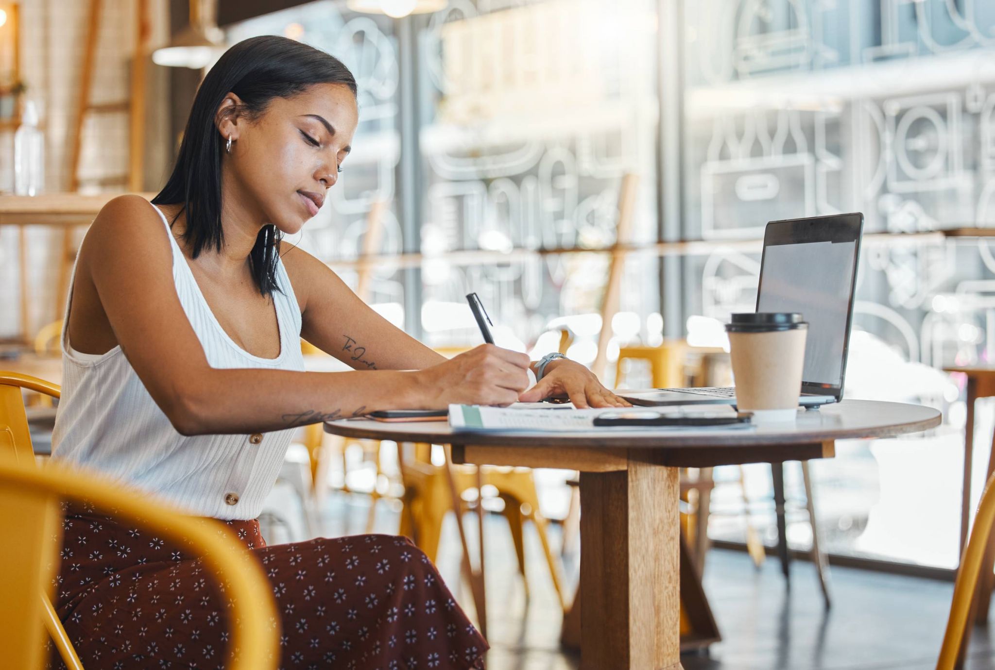Woman writing at a café table with a laptop and coffee cup nearby, seated on a yellow chair.