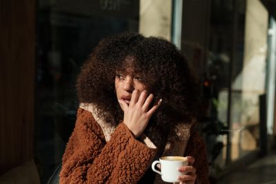 A person with curly hair in a brown coat holds a coffee cup, sitting by a window with sunlight streaming in.
