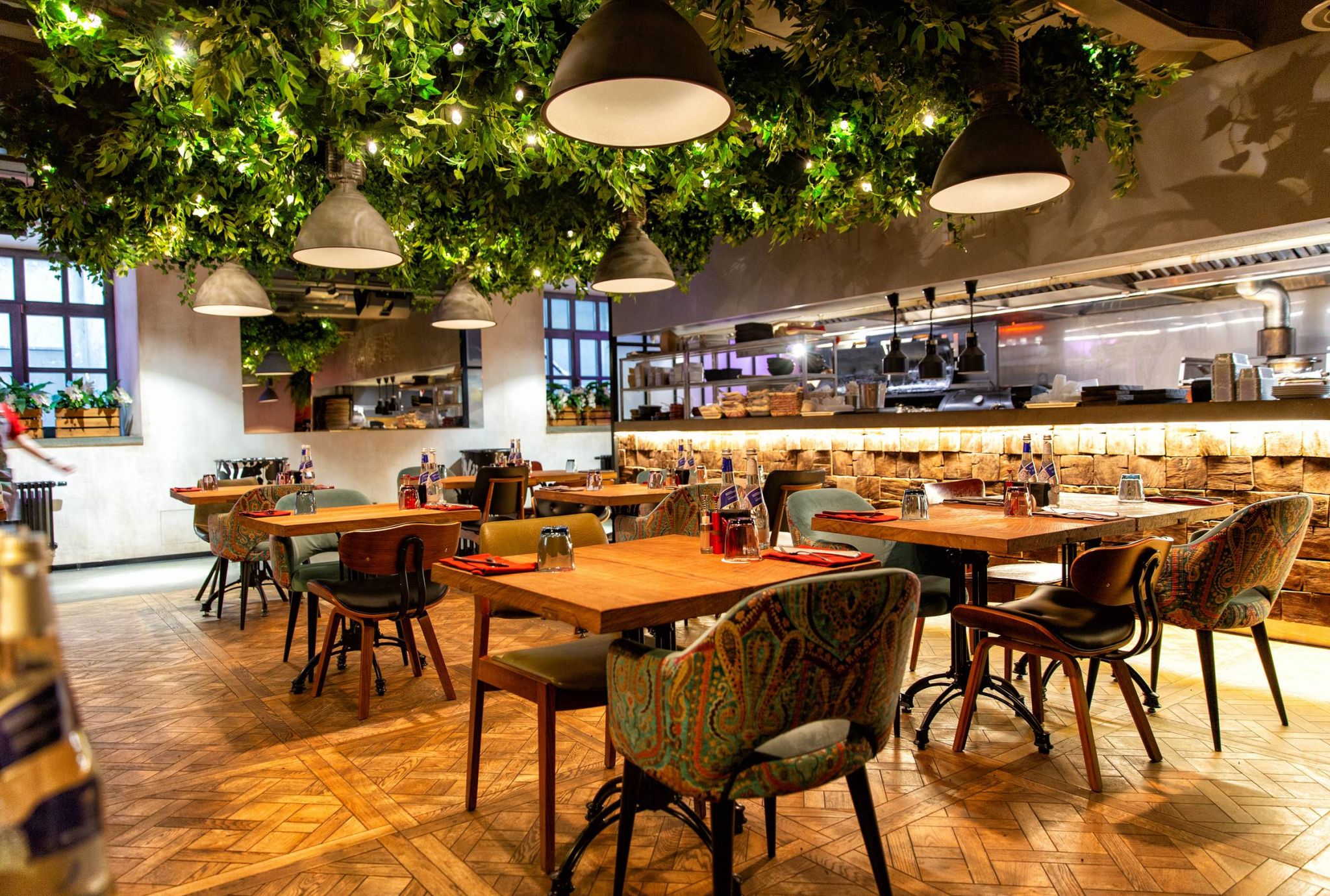 Restaurant interior with wooden tables, patterned chairs, hanging lights, and greenery on the ceiling. Cozy atmosphere.
