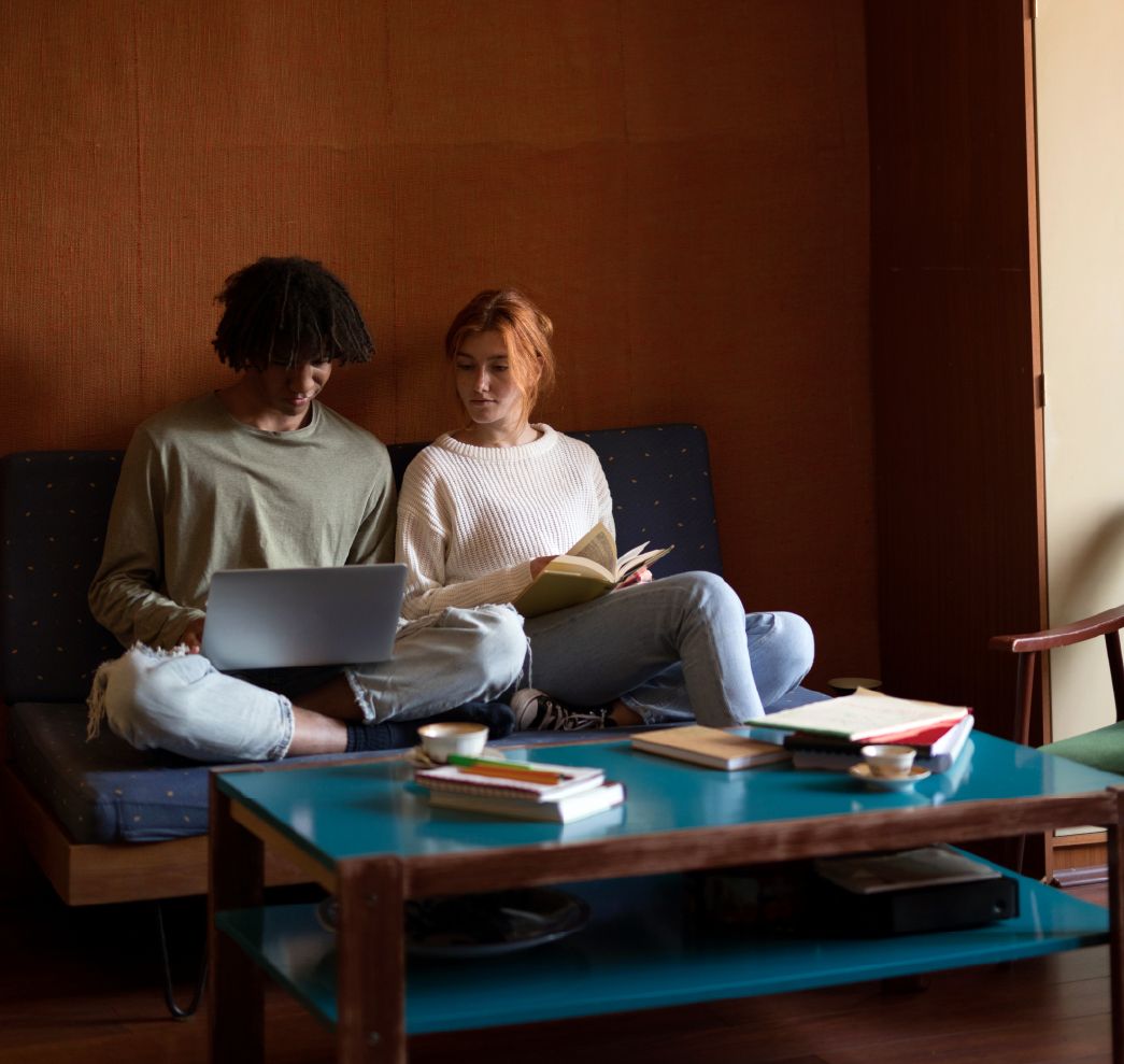 Two people sitting on a couch, using a laptop and reading a book, with a coffee table and books in front of them.