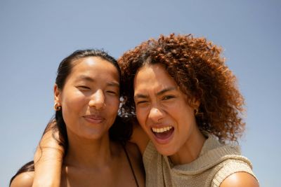 Two people smiling, the person on the right has curly hair and is laughing under a clear blue sky.