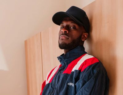 Man in a blue and red jacket leans against a wooden wall, wearing a black hat and a watch.