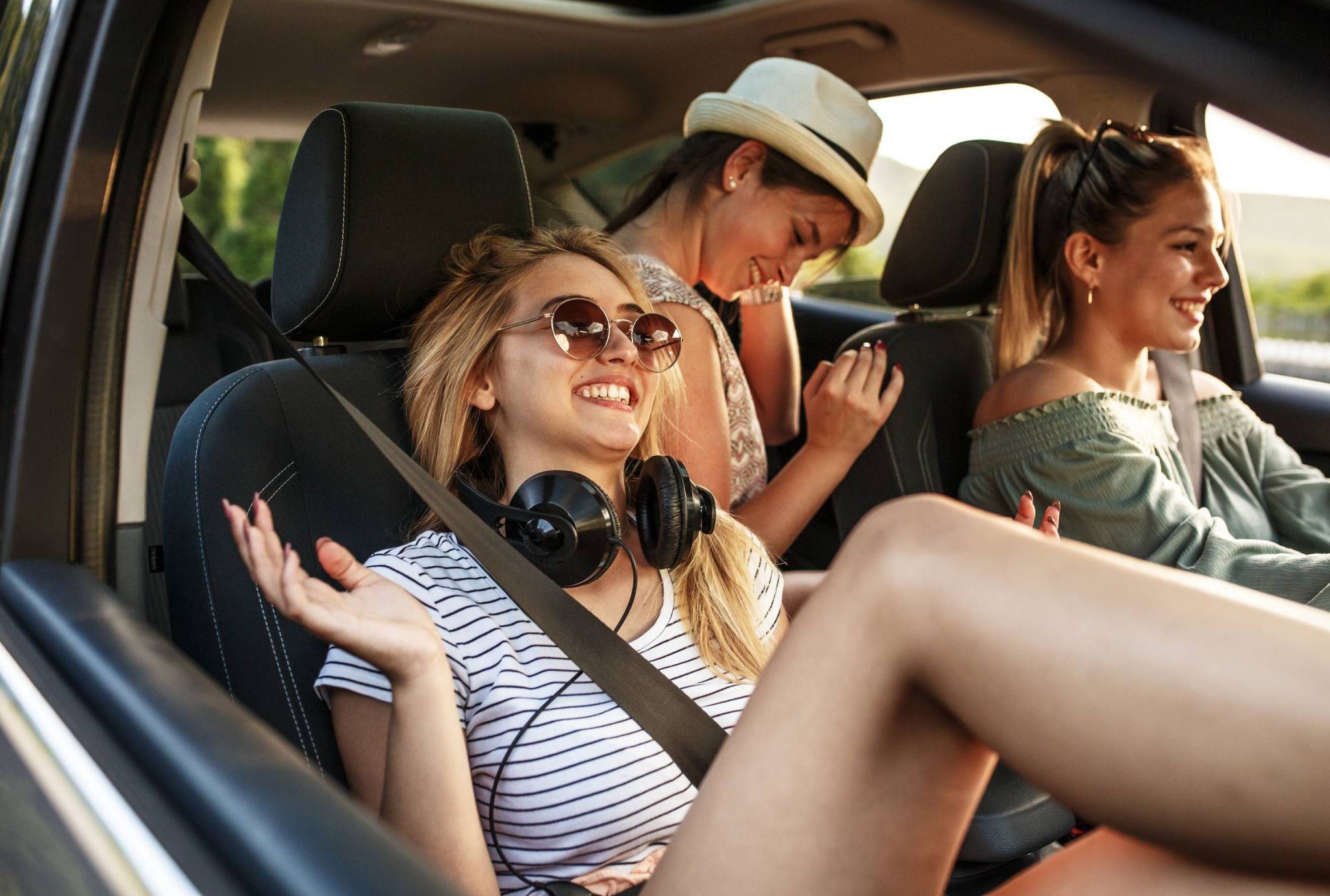 Three young women laughing and relaxing in a car, with one wearing sunglasses and headphones, enjoying a road trip.