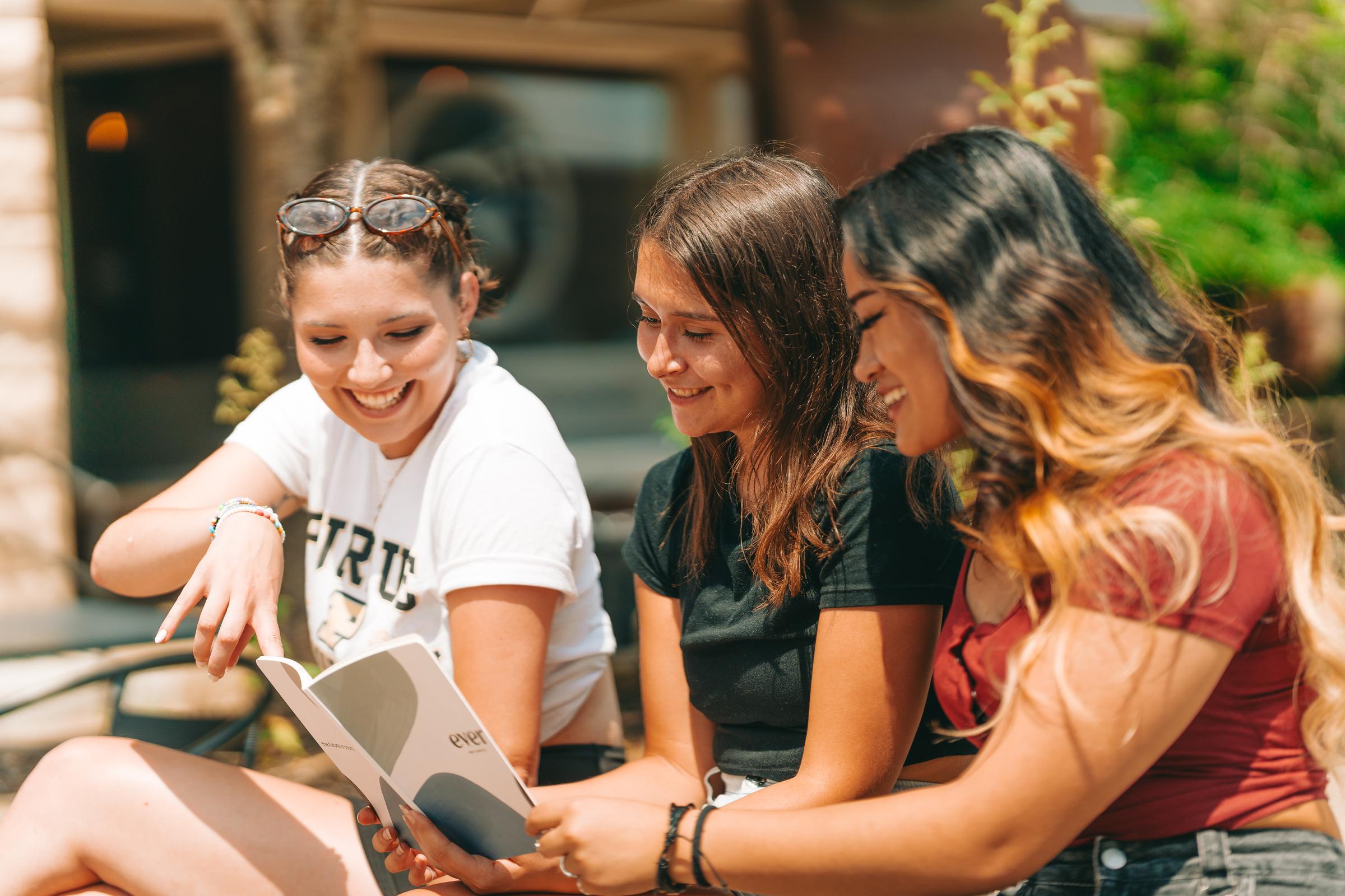 Three female college students sitting outside and looking at a catalog together